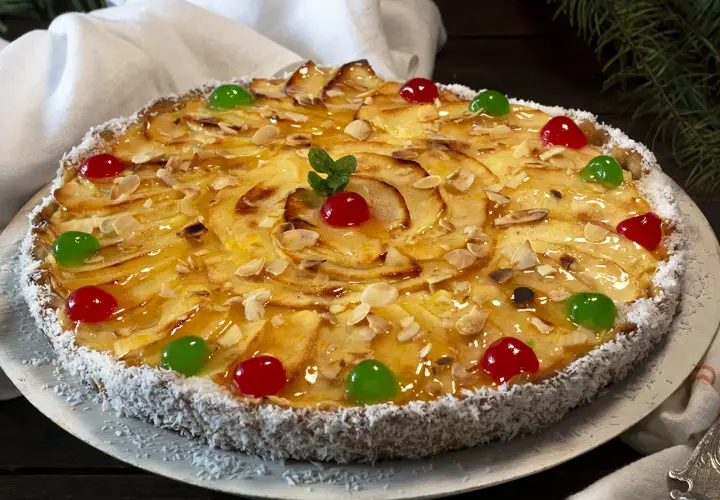 Tarta de manzana con crema pastelera, decorada con almendras laminadas, cerezas rojas y verdes, y coco rallado en el borde.