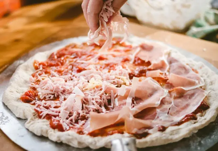 Mano esparciendo queso rallado sobre una masa de pizza casera con jamón y salsa de tomate.
