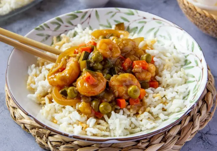 Un plato de arroz blanco cubierto con gambas en salsa agridulce y verduras. Los palillos descansan sobre el borde del cuenco.
