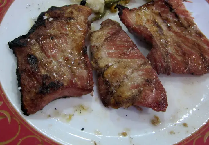 El secreto de cerdo a la parrilla, jugoso y con sal gruesa, servido en un plato blanco con borde rojo.