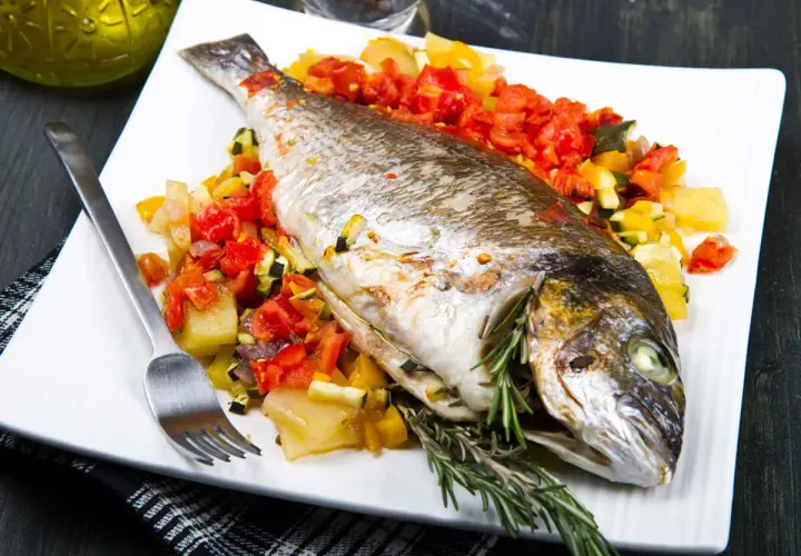 Dorada al horno con verduras asadas, servida en plato blanco con tenedor y romero fresco.