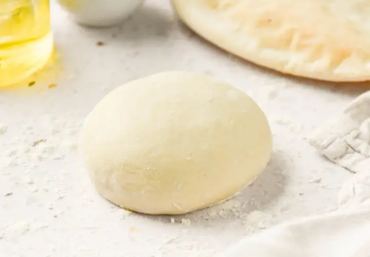 Masa de pizza sin levadura lista para estirar. Harina esparcida, aceite y un trozo de pan plano al fondo.
