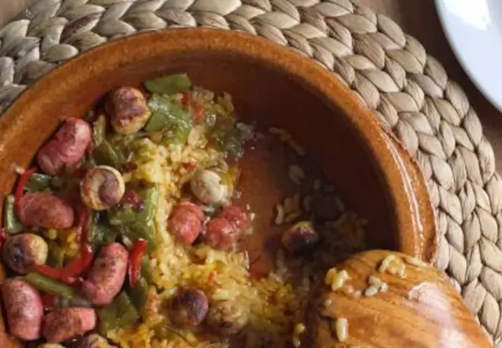 Un plato de barro con arroz, salchichas, pimientos verdes y rojos. Un cucharón de madera reposa en el borde.