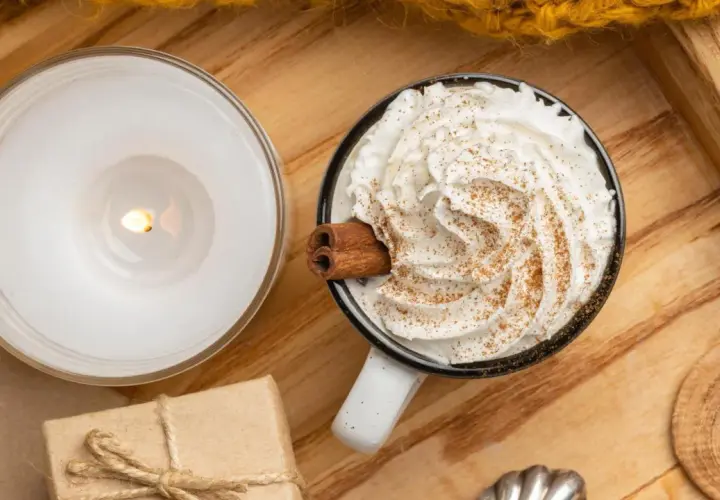 Taza de chocolate caliente con abundante crema de leche, canela y una rama de canela. Una vela encendida y un regalo completan la escena acogedora.