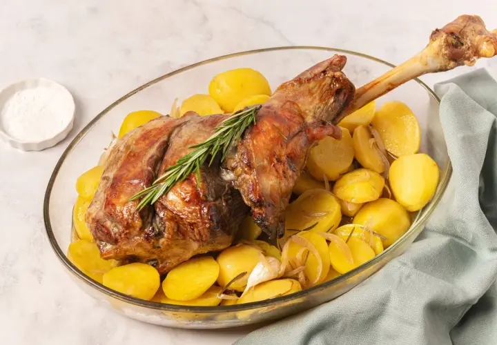 Pierna de cordero al horno dorada y jugosa, acompañada de patatas asadas y romero en una fuente de cristal.