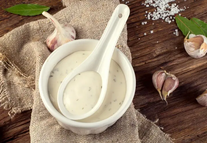 Crema de salsa de ajo en un cuenco blanco con una cuchara, rodeada de ajos y hojas verdes sobre una mesa de madera rústica.