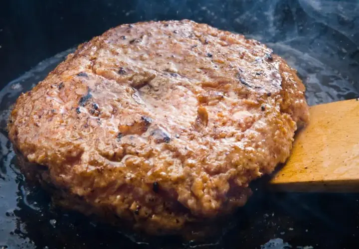 Jugosa hamburguesa de ternera dorándose en sartén, con vapor ascendiendo y una espátula de madera lista para darle la vuelta.
