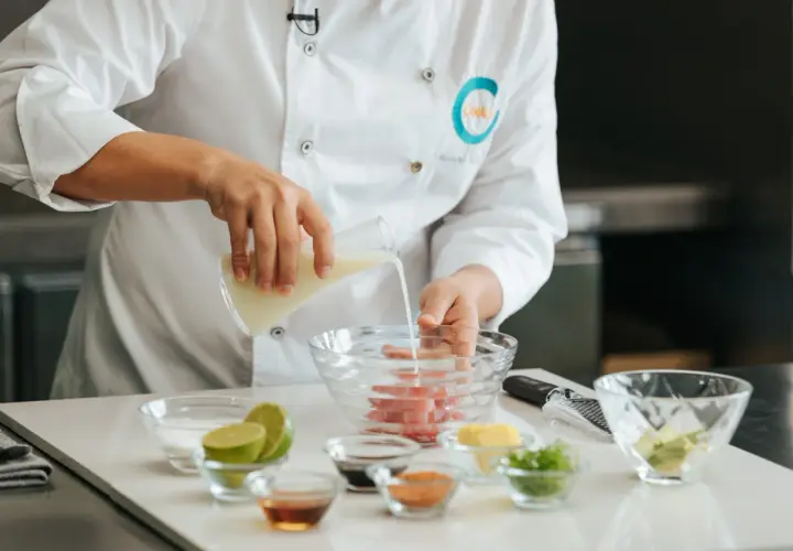 Chef vierte líquido en un bol con pescado, preparando la leche de tigre receta. Ingredientes frescos como limas y aguacate a la vista.