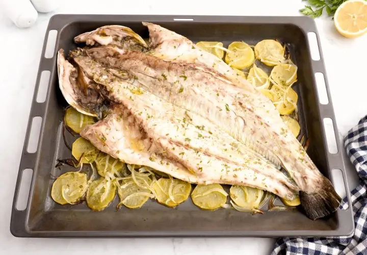 Corvina al horno, entera y dorada, reposa sobre rodajas de patata y cebolla en una bandeja.