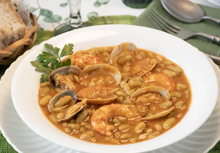 Plato de verdinas con almejas y gambas, servido en un cuenco blanco con perejil. Acompañado de pan en una cesta.