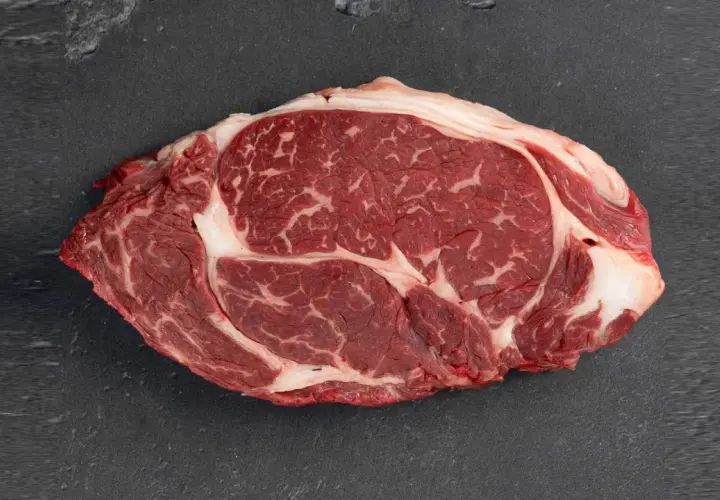 Un corte de aguja de ternera cruda, con marmoleo de grasa blanca entre la carne roja, sobre una superficie oscura.