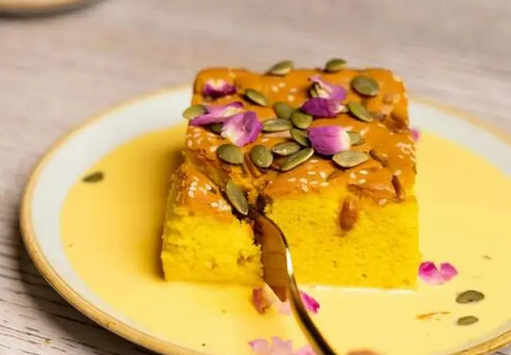 Un trozo de bizcocho de calabaza, decorado con semillas de calabaza y pétalos de rosa, servido en una crema amarilla.