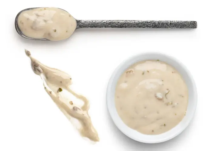 Cuchara con salsa de champiñones cremosa, un chorrito de la misma salsa y un cuenco blanco lleno de esta deliciosa salsa.