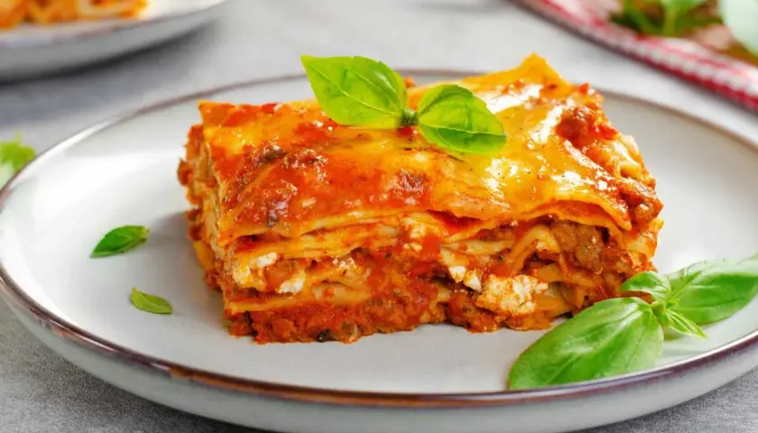 Una porción de lasaña de carne, con capas de pasta, salsa y queso, decorada con hojas de albahaca fresca.
