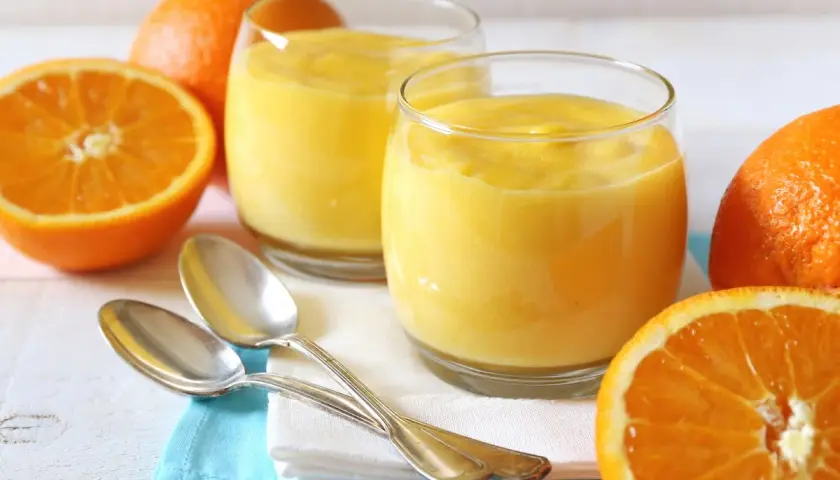 Dos vasos de crema de naranja, naranjas frescas y cucharas plateadas sobre servilletas.