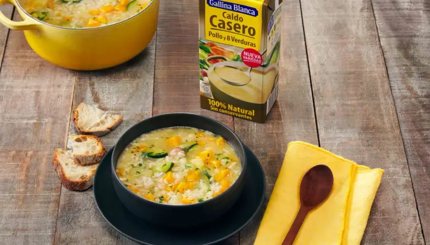 Un tazón humeante de sopa de arroz con calabacín y zanahoria, junto a pan rústico y un cartón de caldo Gallina Blanca.