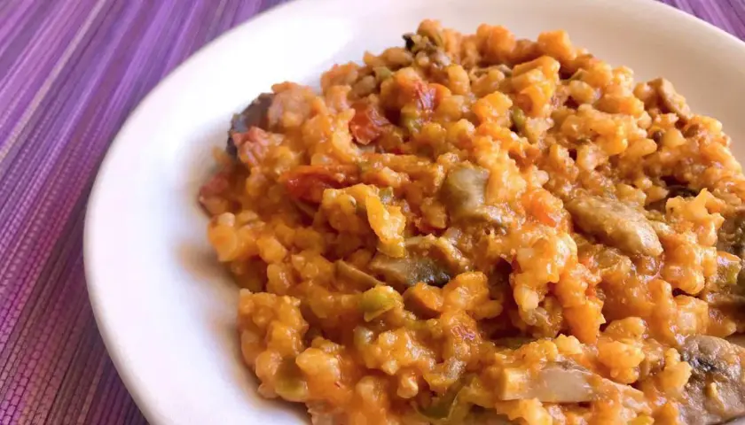 Un cremoso risotto de calabaza con trozos de champiñones y pimientos, servido en un plato blanco sobre un mantel morado.