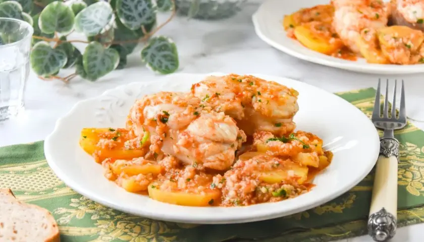 Delicioso rape con patatas en salsa de tomate y ajo, servido en un plato blanco con un tenedor y pan.