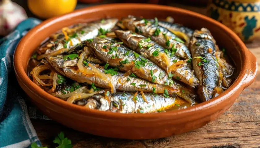 Sardinas en escabeche, tiernas y jugosas, reposan en una cazuela de barro, adornadas con perejil fresco y cebolla caramelizada.