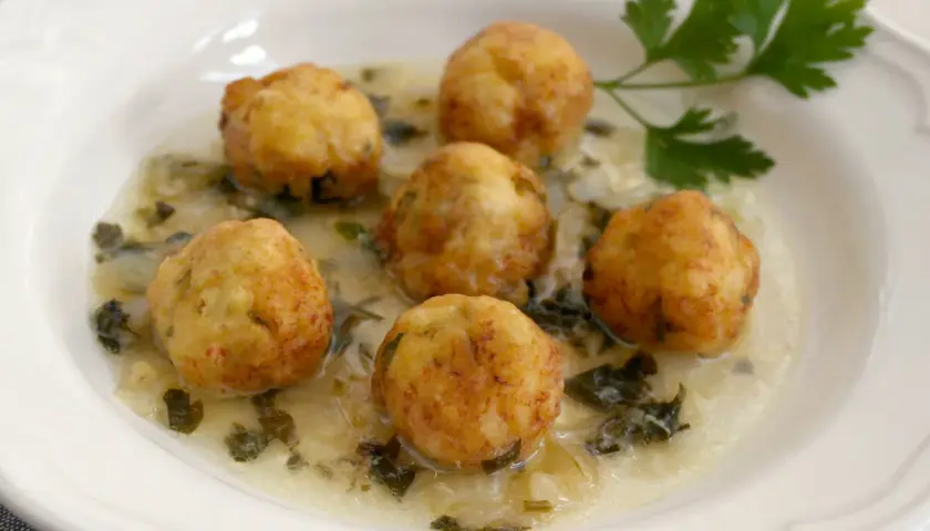 Seis albóndigas de merluza doradas flotan en una cremosa salsa verde con perejil fresco.