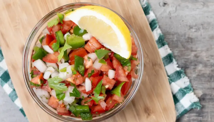 Un tazón de pico de gallo fresco, con tomate, cebolla, pimiento verde y cilantro, adornado con una rodaja de limón.