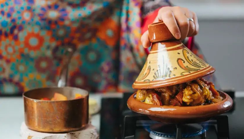 Una mano levanta la tapa de un tajín, revelando un guiso humeante. Descubre qué es el tajín, un plato marroquí.