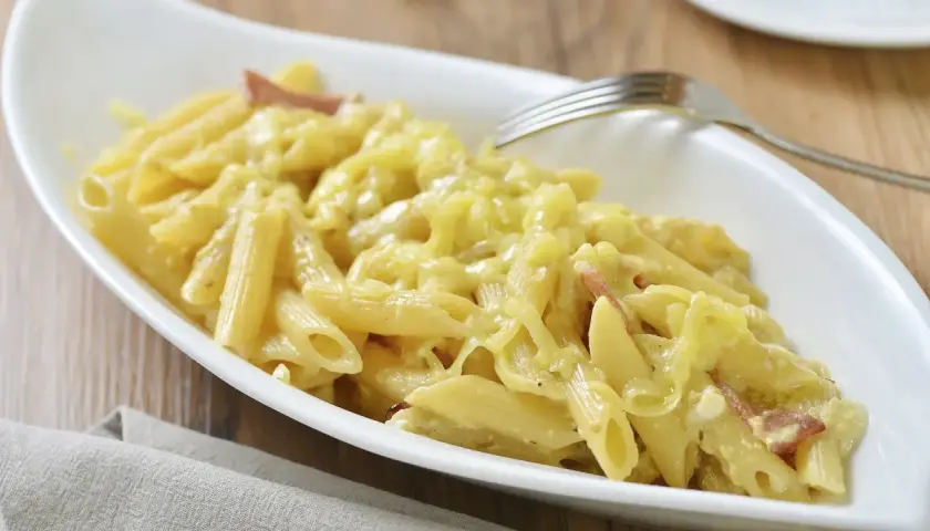 Un plato blanco ovalado lleno de macarrones con queso cremosos y trozos de tocino. Un tenedor plateado descansa sobre el borde del plato.