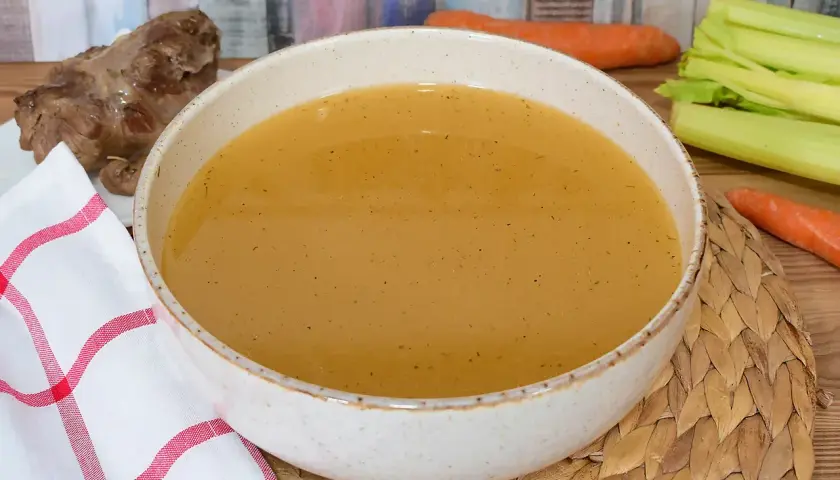 Un tazón de caldo de carne humeante, con un trozo de carne y verduras frescas al lado.