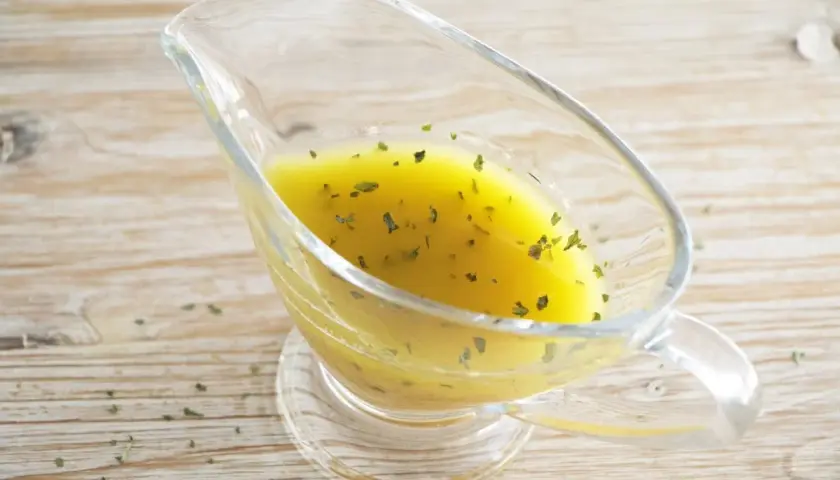 Vinagreta de mostaza dorada con hierbas verdes picadas, servida en una salsera de cristal sobre una mesa de madera.