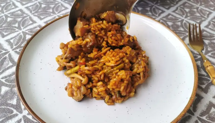 Un cucharón sirve arroz con secreto, un plato sabroso con carne y verduras, en un plato blanco.