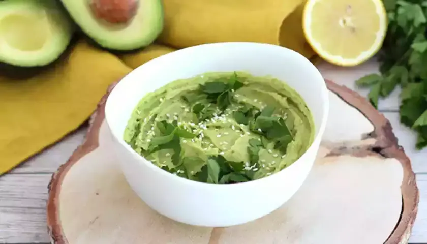 Un cremoso hummus de aguacate, decorado con perejil fresco y semillas de sésamo, junto a aguacates cortados y un limón.