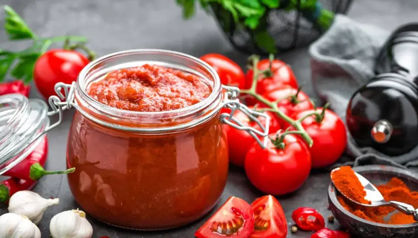 Frasco de vidrio lleno de vibrante salsa de tomate casera, rodeado de tomates frescos, ajos, chiles y especias.