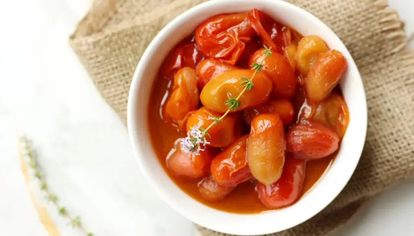 Un tazón blanco lleno de tomates cherry confitados, con una ramita de tomillo y una flor morada.
