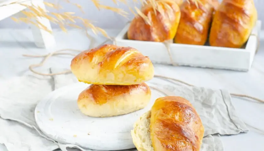 Dos panes de leche dorados apilados, uno abierto, con más panes en una bandeja al fondo.