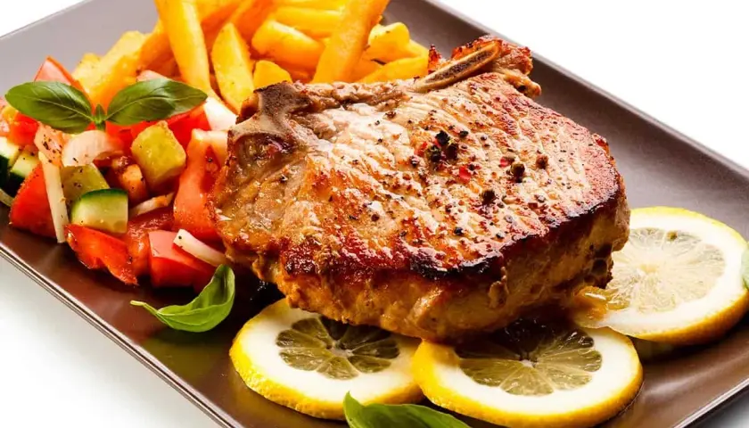 Chuletas de cerdo al horno jugosas, acompañadas de papas fritas crujientes, ensalada fresca y rodajas de limón.