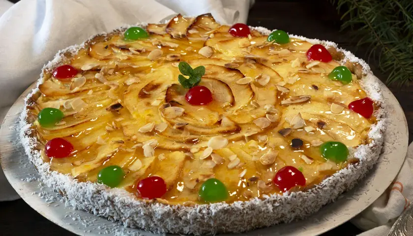 Tarta de manzana con crema pastelera, decorada con almendras laminadas, cerezas rojas y verdes, y coco rallado en el borde.