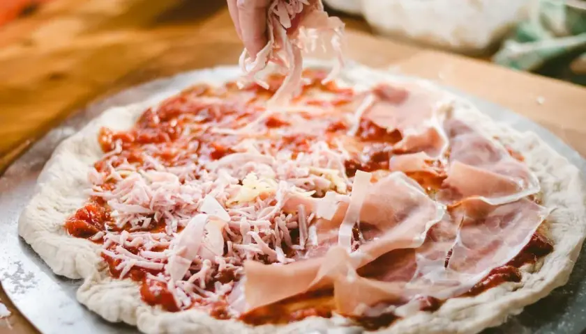 Mano esparciendo queso rallado sobre una masa de pizza casera con jamón y salsa de tomate.