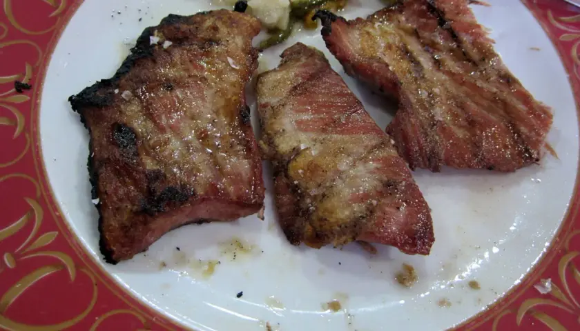 El secreto de cerdo a la parrilla, jugoso y con sal gruesa, servido en un plato blanco con borde rojo.
