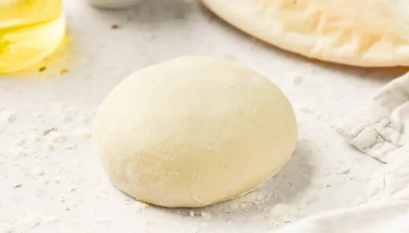 Masa de pizza sin levadura lista para estirar. Harina esparcida, aceite y un trozo de pan plano al fondo.
