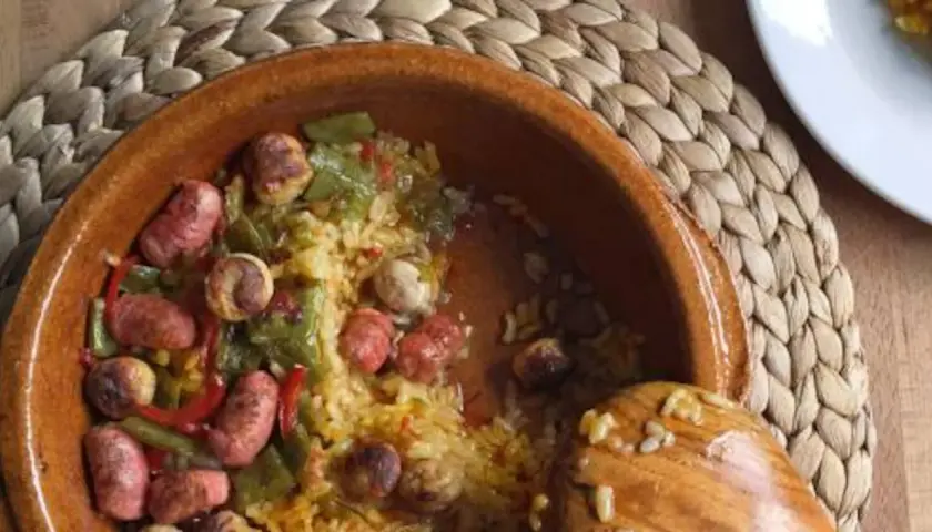 Un plato de barro con arroz, salchichas, pimientos verdes y rojos. Un cucharón de madera reposa en el borde.