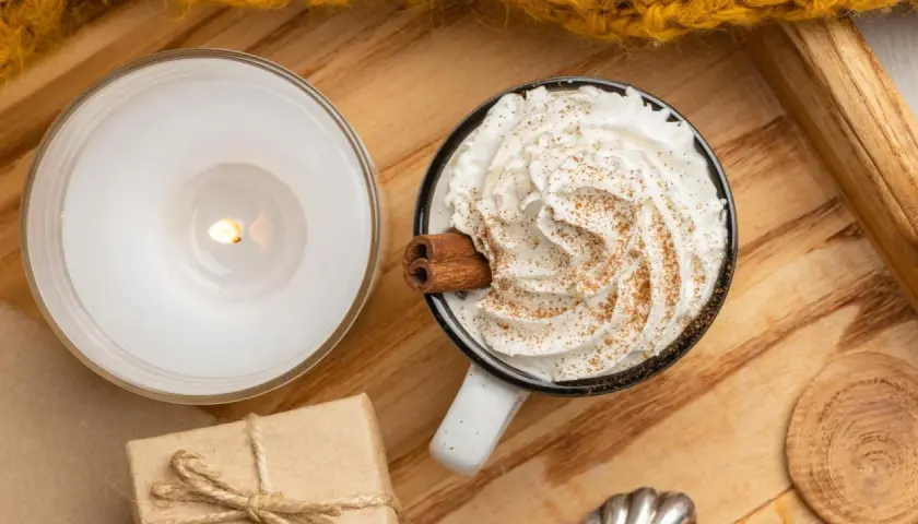 Taza de chocolate caliente con abundante crema de leche, canela y una rama de canela. Una vela encendida y un regalo completan la escena acogedora.