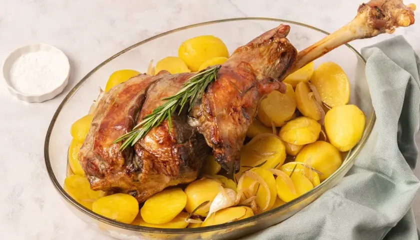 Pierna de cordero al horno dorada y jugosa, acompañada de patatas asadas y romero en una fuente de cristal.