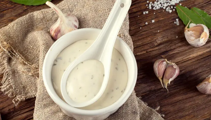 Crema de salsa de ajo en un cuenco blanco con una cuchara, rodeada de ajos y hojas verdes sobre una mesa de madera rústica.