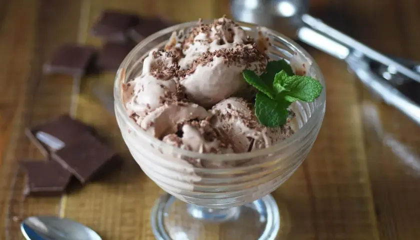 Delicioso helado de chocolate casero en copa, adornado con virutas de chocolate y una hoja de menta. A su lado, trozos de chocolate negro y una cuchara.
