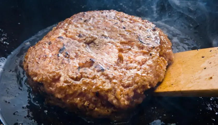 Jugosa hamburguesa de ternera dorándose en sartén, con vapor ascendiendo y una espátula de madera lista para darle la vuelta.