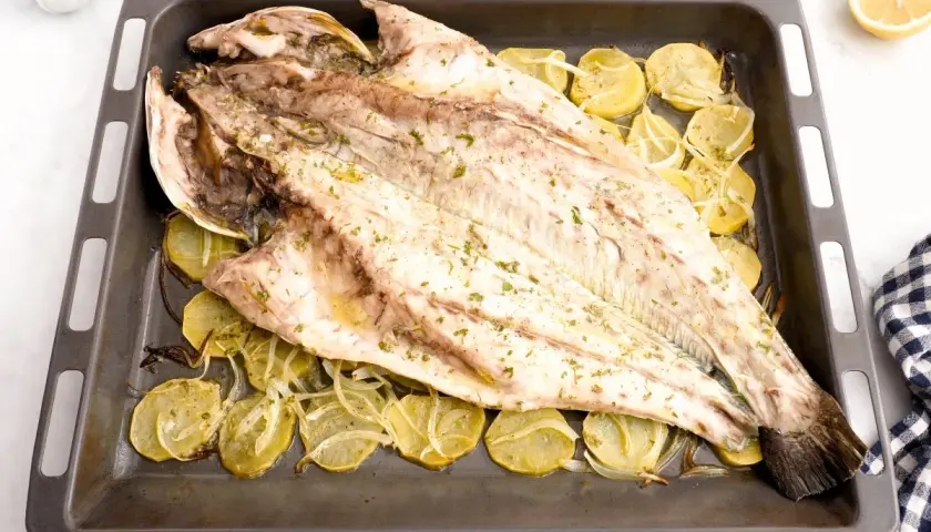 Corvina al horno, entera y dorada, reposa sobre rodajas de patata y cebolla en una bandeja.