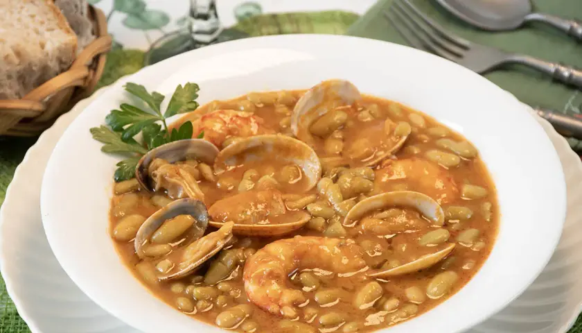 Plato de verdinas con almejas y gambas, servido en un cuenco blanco con perejil. Acompañado de pan en una cesta.