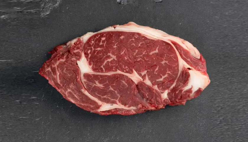 Un corte de aguja de ternera cruda, con marmoleo de grasa blanca entre la carne roja, sobre una superficie oscura.