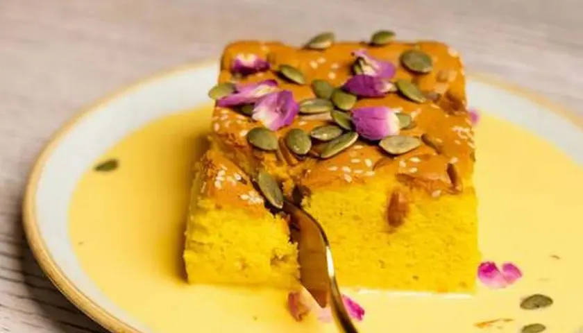 Un trozo de bizcocho de calabaza, decorado con semillas de calabaza y pétalos de rosa, servido en una crema amarilla.