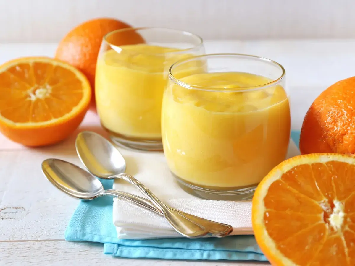 Dos vasos de crema de naranja, naranjas frescas y cucharas plateadas sobre servilletas.